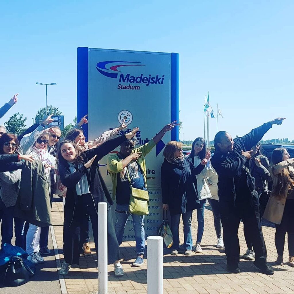 Grupo de personas posando con entusiasmo frente al cartel del Madejski Stadium, haciendo el gesto de "dabbing" al aire libre.